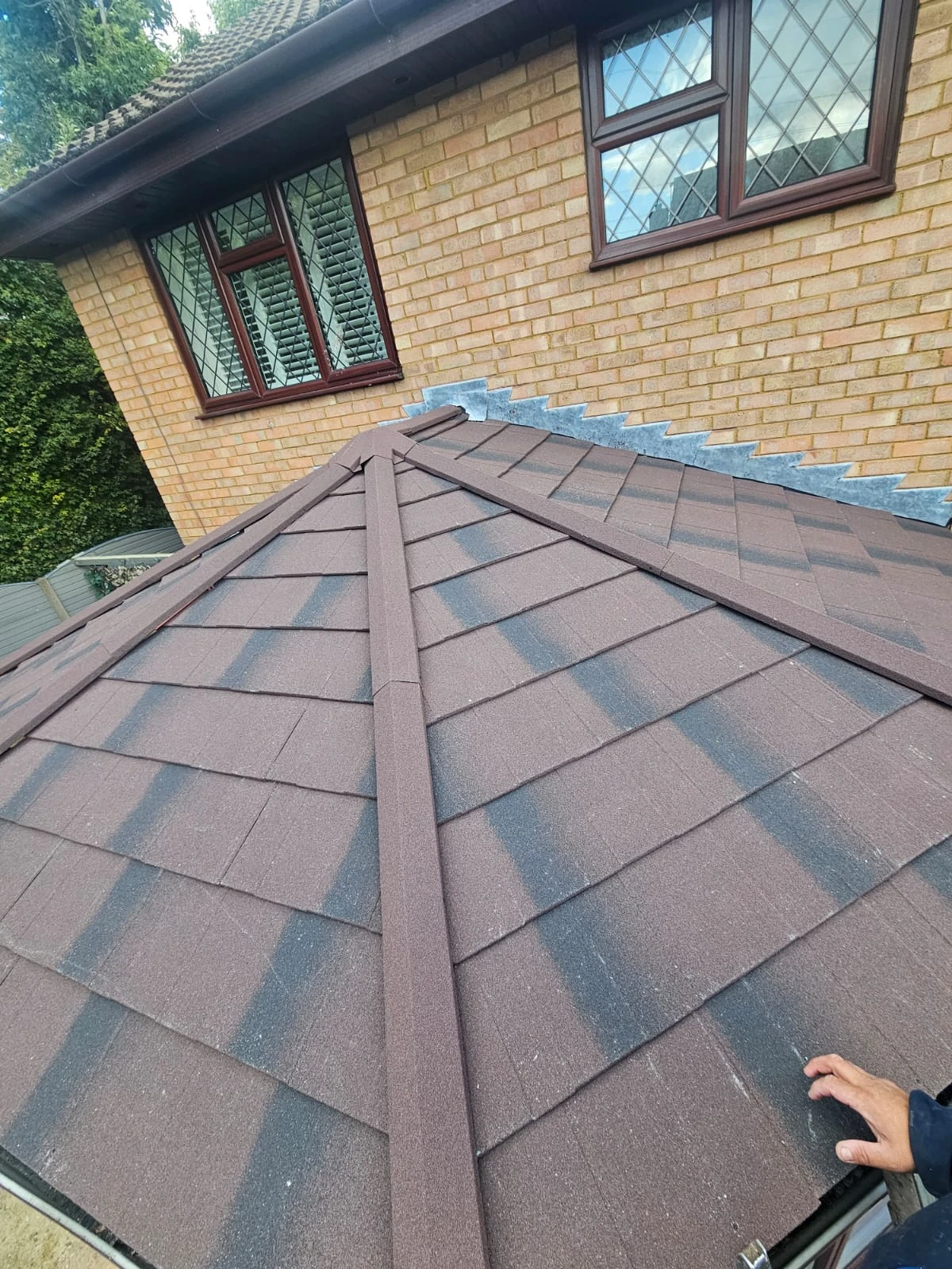 A close-up view of a brown, textured roof with black streaks, and a hand partially visible on the edge. A brick wall is in the background.
